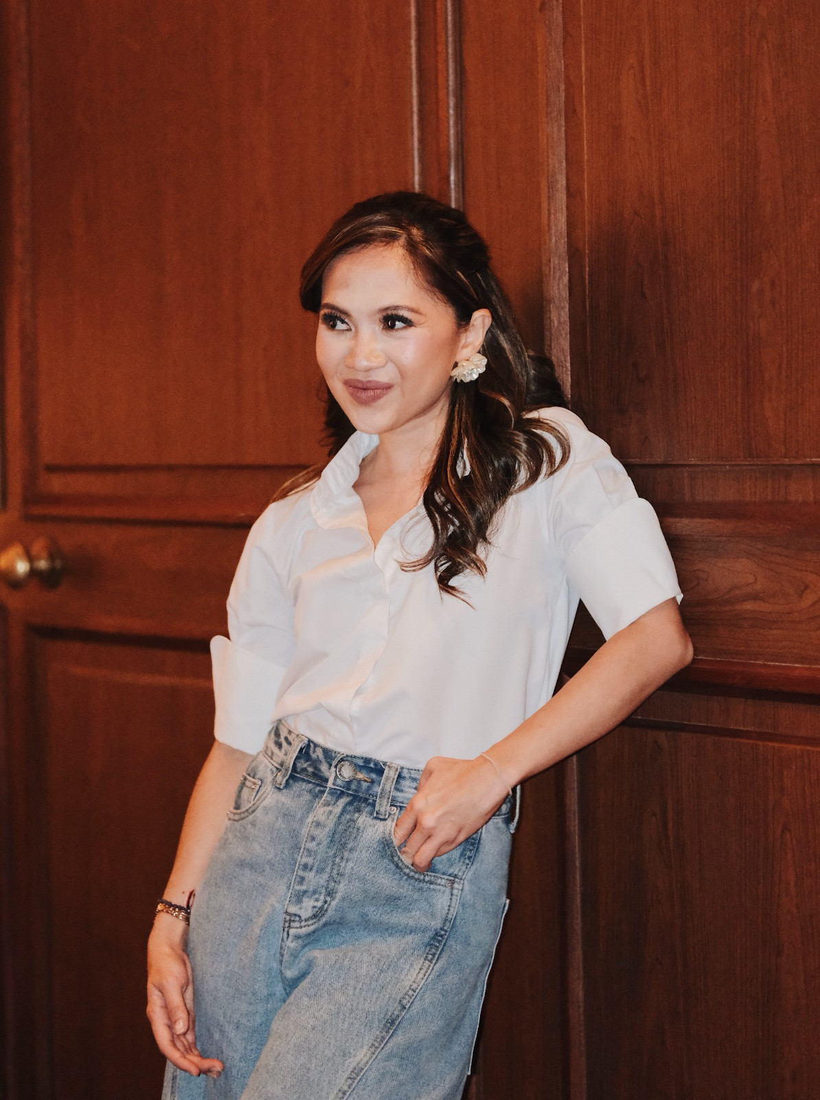 A woman in a white button-up shirt and light-wash jeans offers a thoughtful smile.