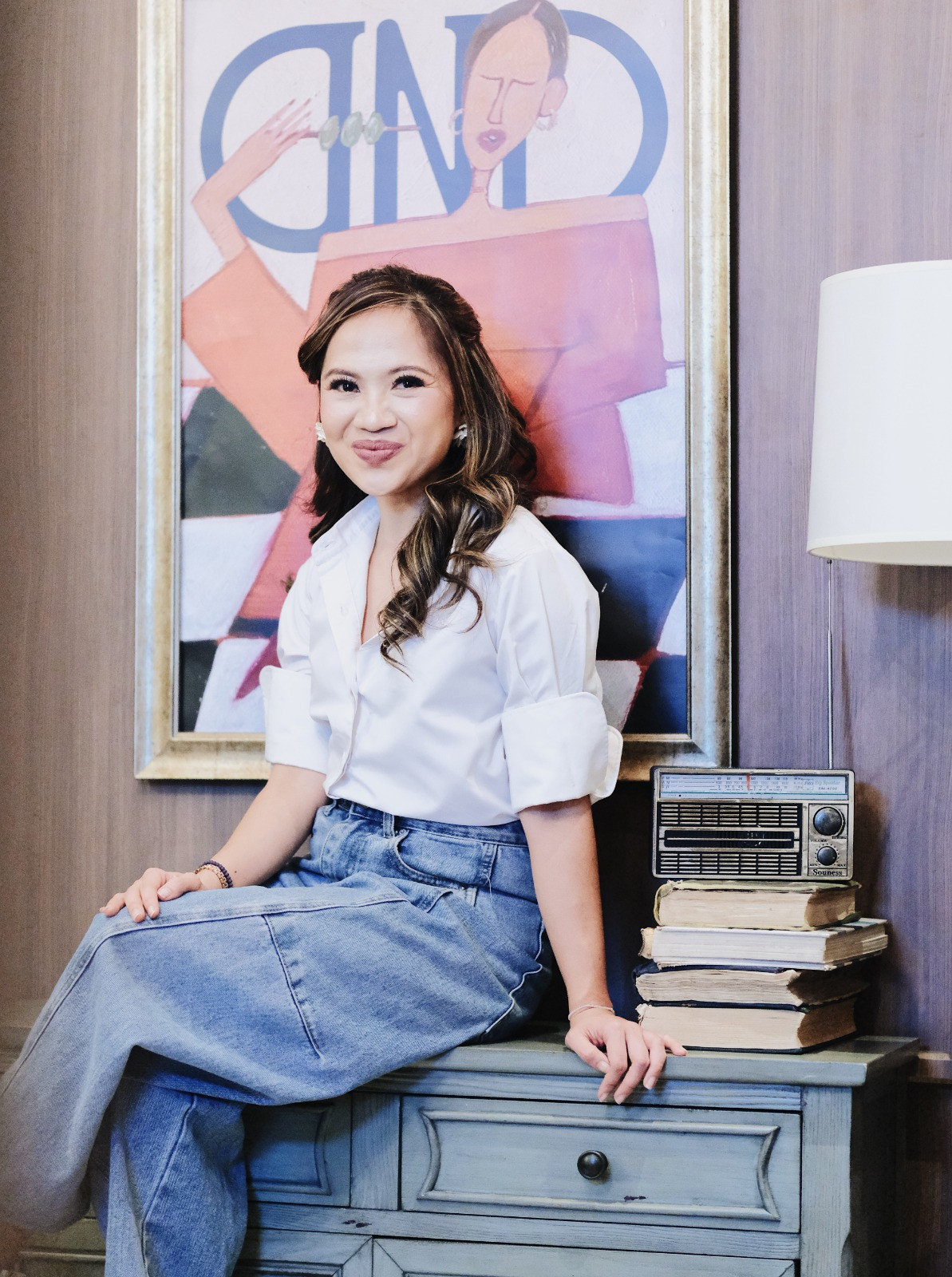 A woman smiles from a well-appointed interior, featuring a console table, vintage radio, and framed artwork.