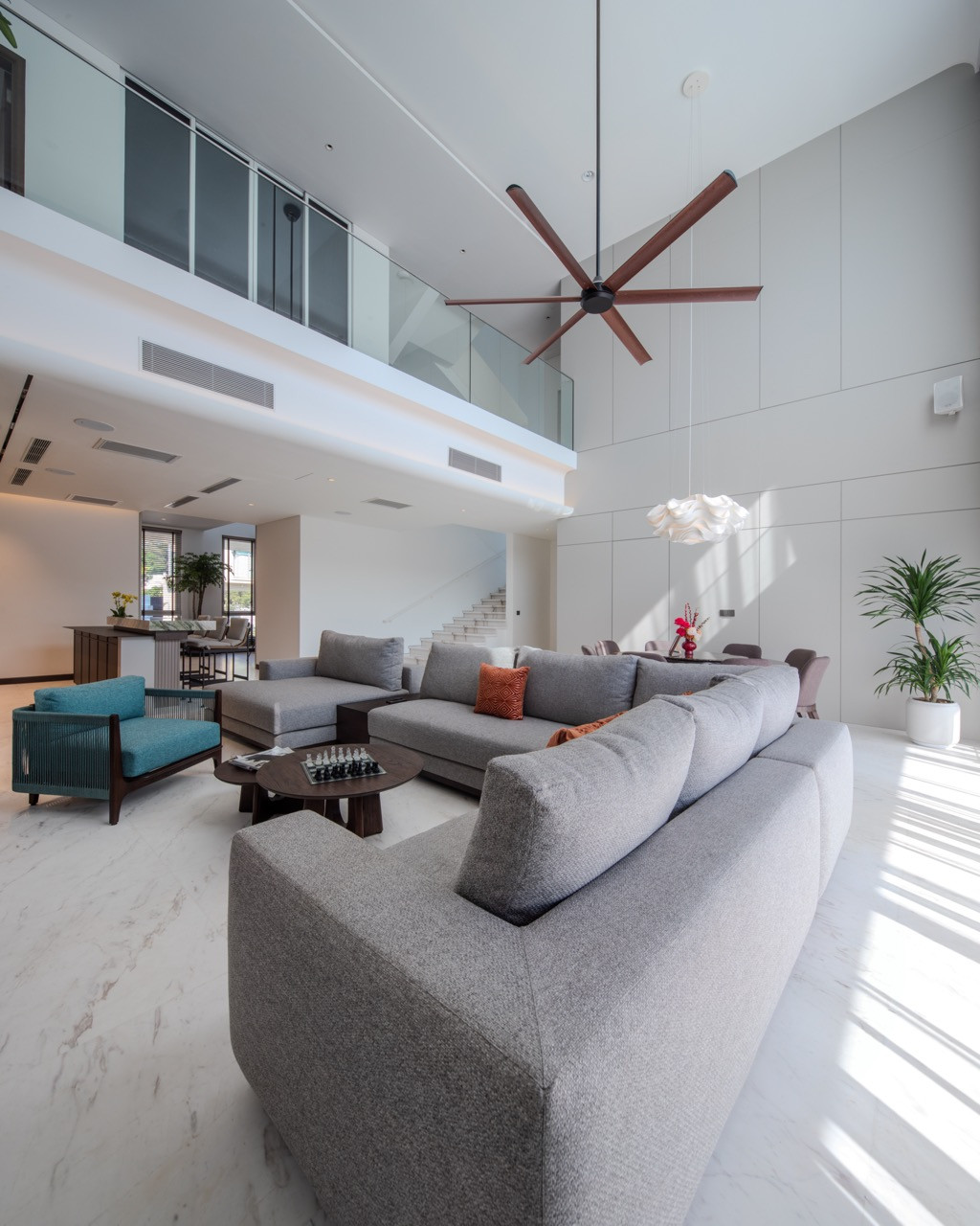 A view across the double-volume living hall toward the staircase and the mezzanine level above, with a glazed balustrade marking the edge of the upper floor