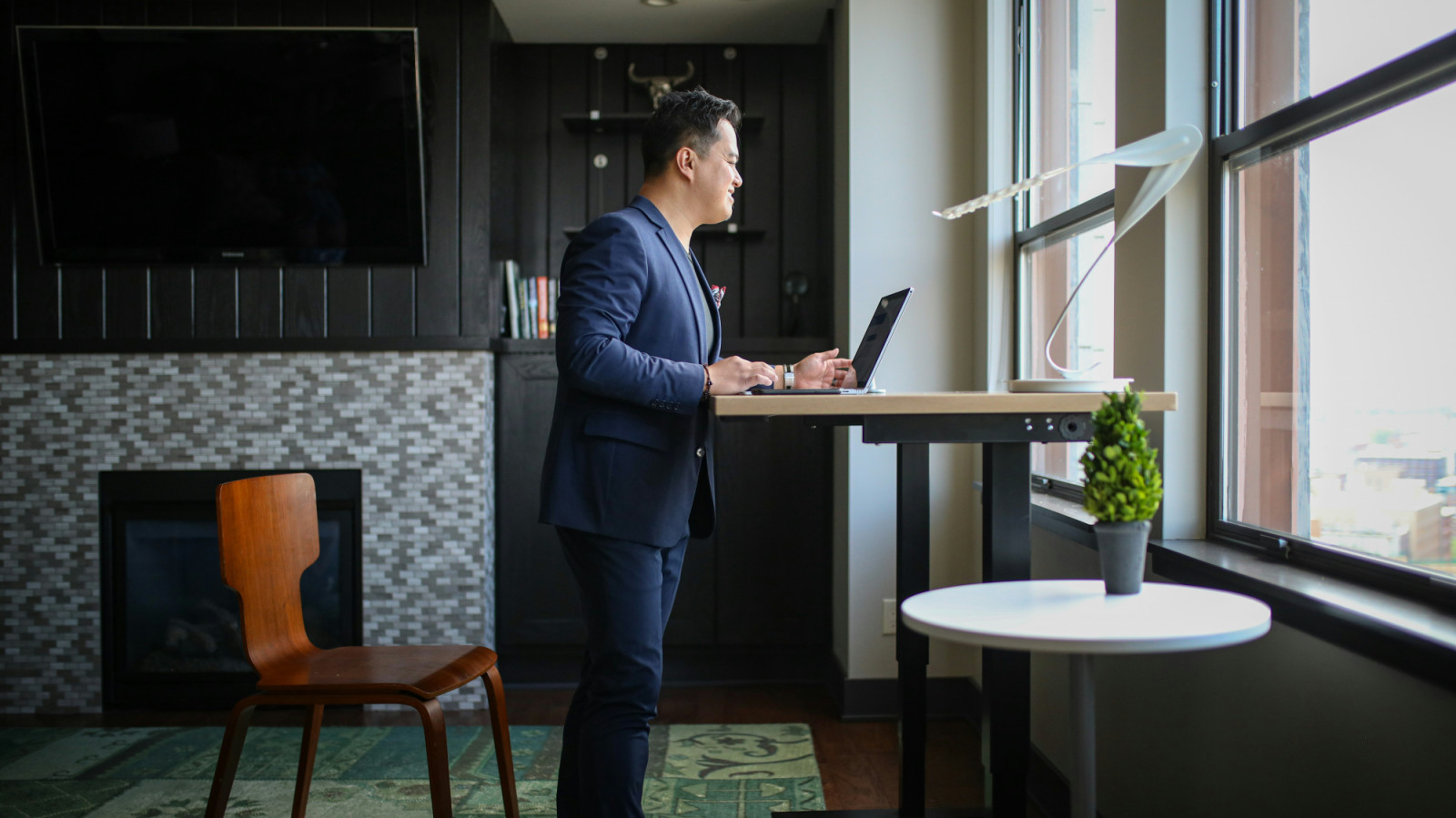 A man works on his laptop at a standing desk in a modern office, gazing out the window.