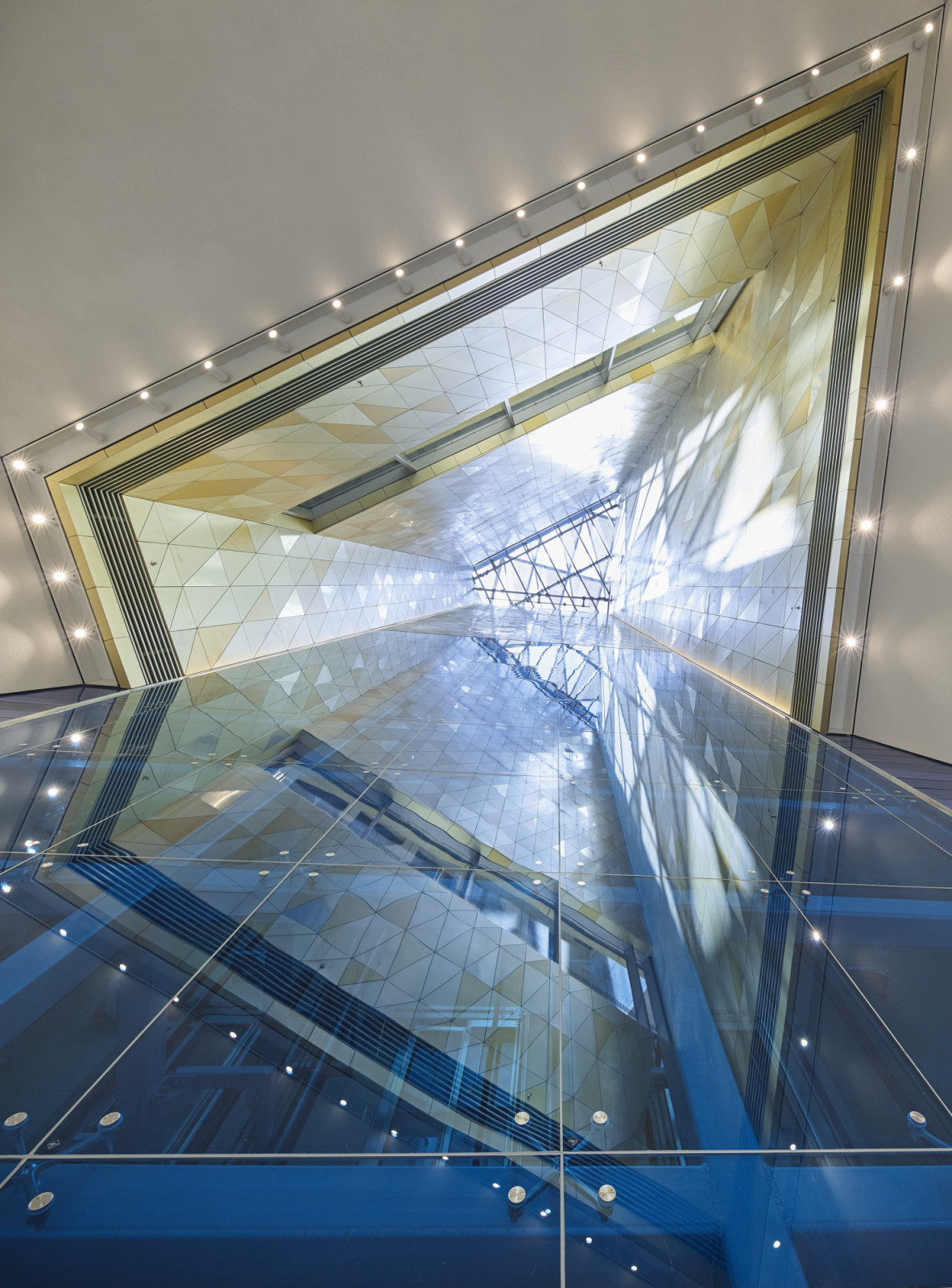 Seen from below a glass floor, a triangular light well lined with white and gold faceted panels rises towards a skylight (Photo: Hufton+Crow)