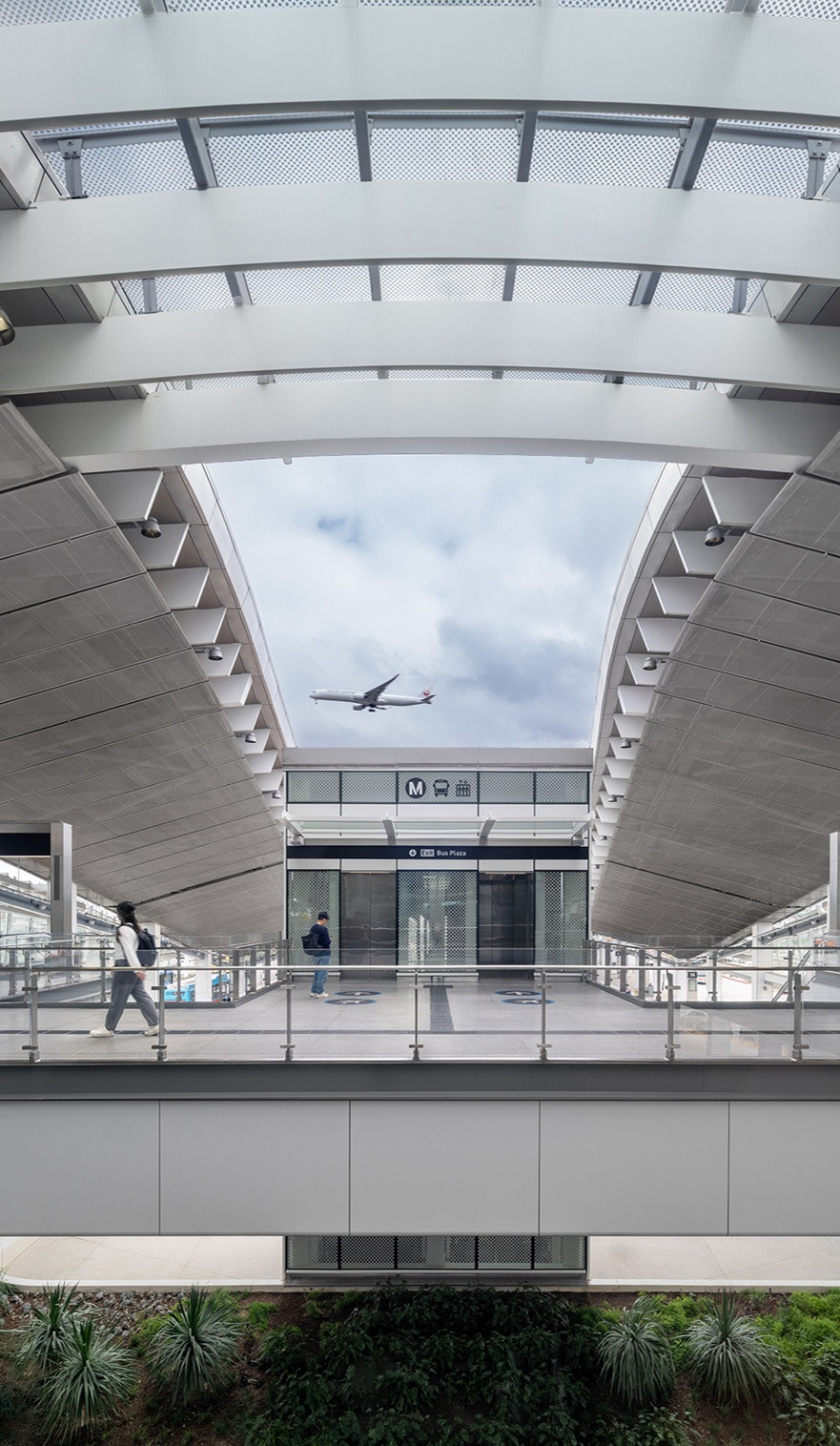 A plane passes overhead as passengers cross the upper concourse of the LAX/Metro Transit Center in Los Angeles, beneath sweeping canopies and a ribbed glass roof(Photo: Jason O’Rear)