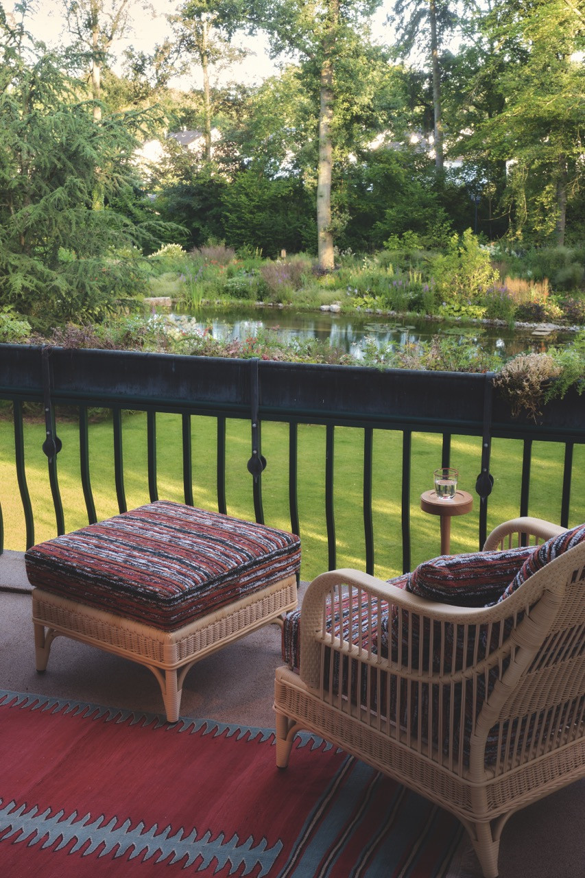 Wicker armchairs on the terrace look out over the natural swimming pond and garden beyond