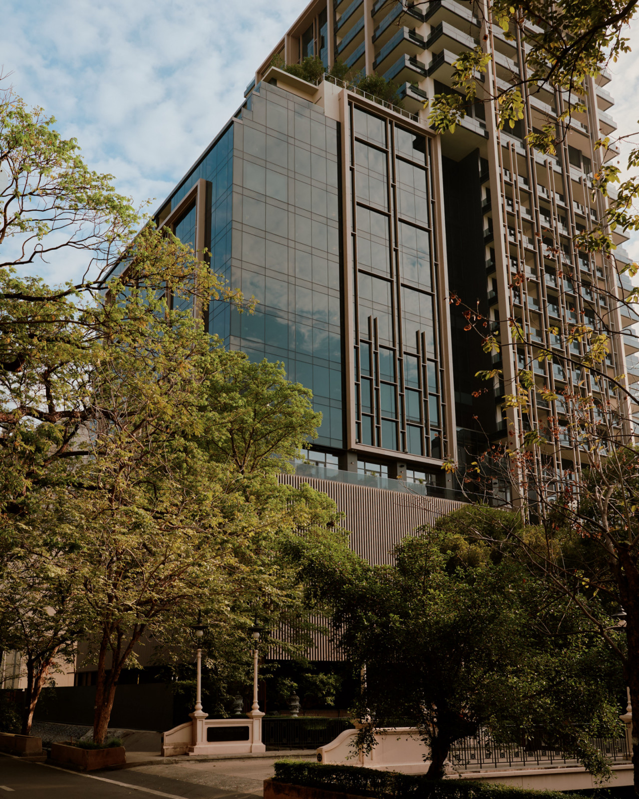 Aman Nai Lert Bangkok rises behind the mature trees of Nai Lert Park, with a dark glass facade above a ribbed podium (Photo: Aman)