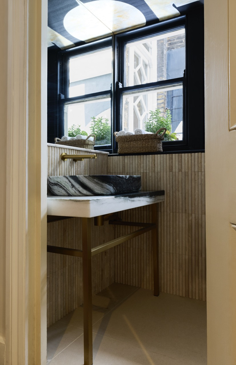The cloakroom, glimpsed from the doorway, where a marble vessel basin on a brass-framed stand sits against fluted travertine tiles beneath a graphic ceiling paper