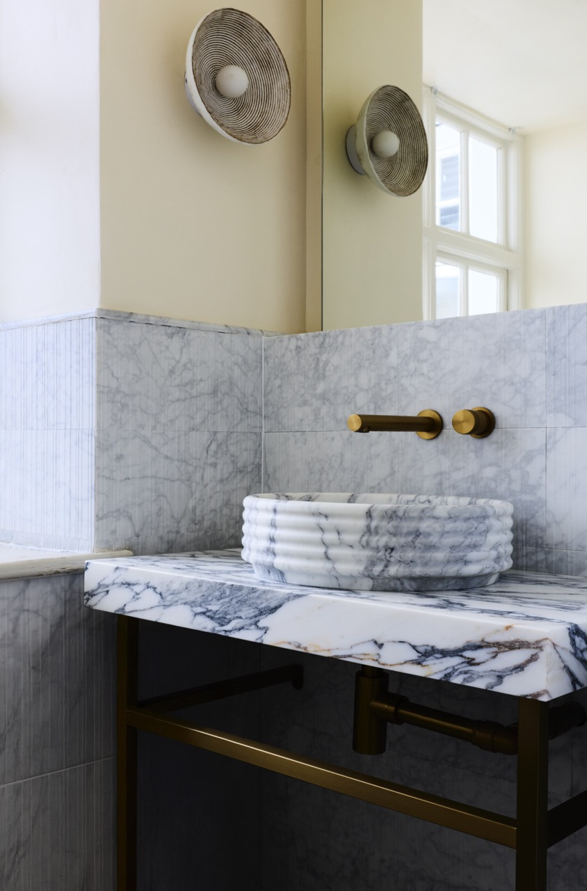 A ribbed marble basin from Lusso on a brass-framed vanity, with brushed brass taps and textured ceramic wall lights