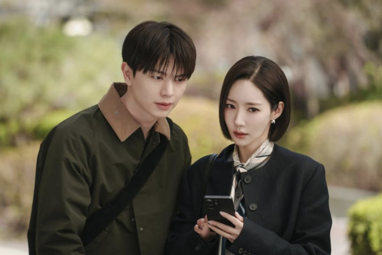 Two actors from a K-drama series share a scene, engaging with a smartphone outdoors.