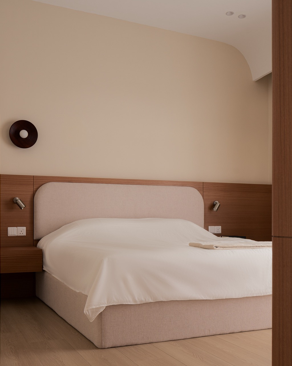 In the bedroom, an upholstered bed with a rounded headboard sits against a laminate panel fitted with reading lights, beneath a curved ceiling edge