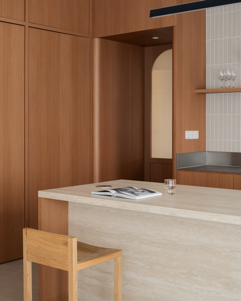 A natural wood stool by the studio sits at the sintered stone island, near the arched reeded-glass door to the kitchen