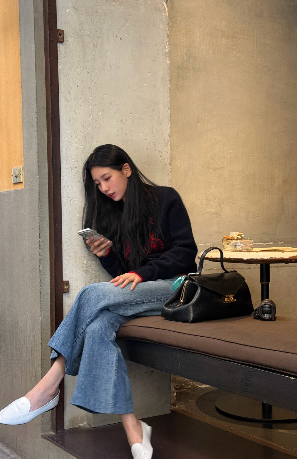 Bae Suzy, the actress and singer, checks her phone while seated, a sophisticated black handbag resting nearby.