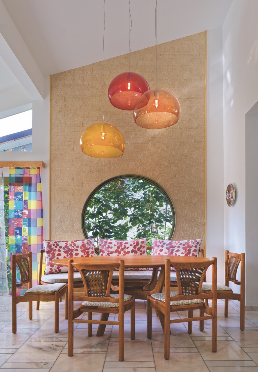 The dining nook’s cluster of red, amber and green glass pendants glows above a table set beside a circular window framing the garden