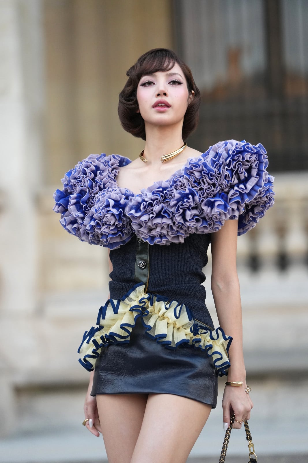 Lisa, Louis Vuitton Global Ambassador, wears an off-the-shoulder ruffled top and leather mini skirt.