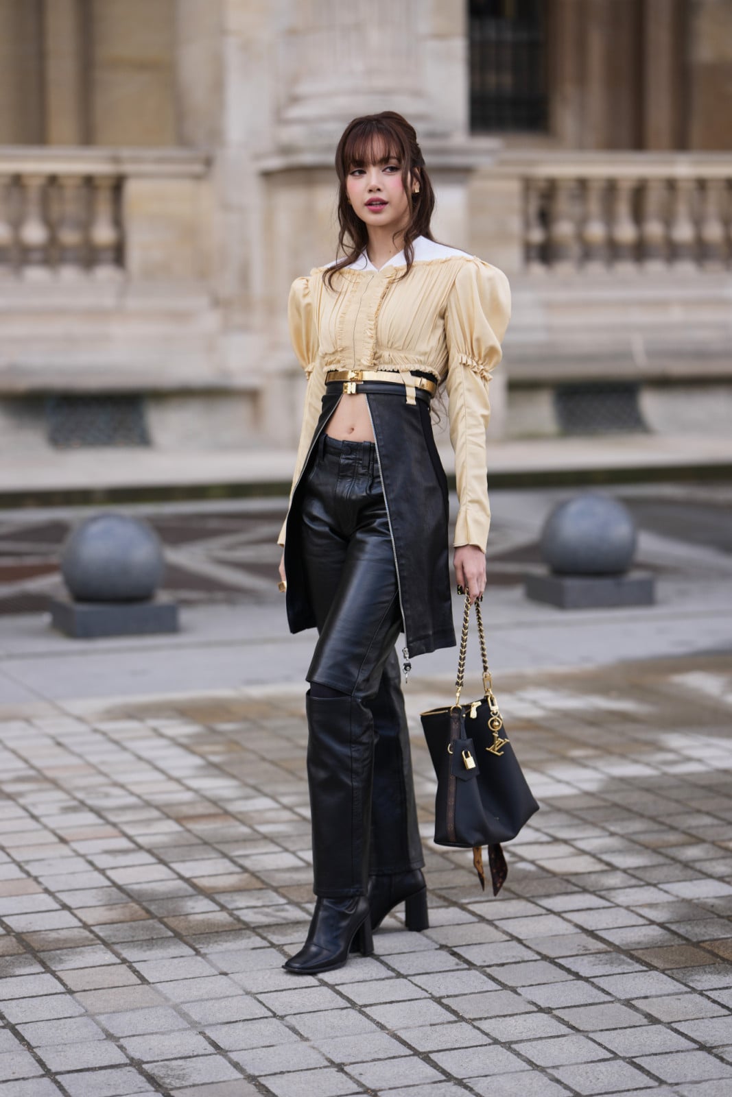 PARIS, FRANCE - MARCH 10: Lisa Manobal wears dark brown long hair with bangs, a beige satin puff sleeve cropped gathered and pleated blouse shirt with a white pointed collar, Louis Vuitton double belts in gold and black leather, black leather straight leg pants, a black leather bag, black leather ankle boots with square toe, outside Louis Vuitton, during Paris Fashion Week - Womenswear Fall/Winter 2026/2027, on March 10, 2026 in Paris, France (Photo by Edward Berthelot/Getty Images)