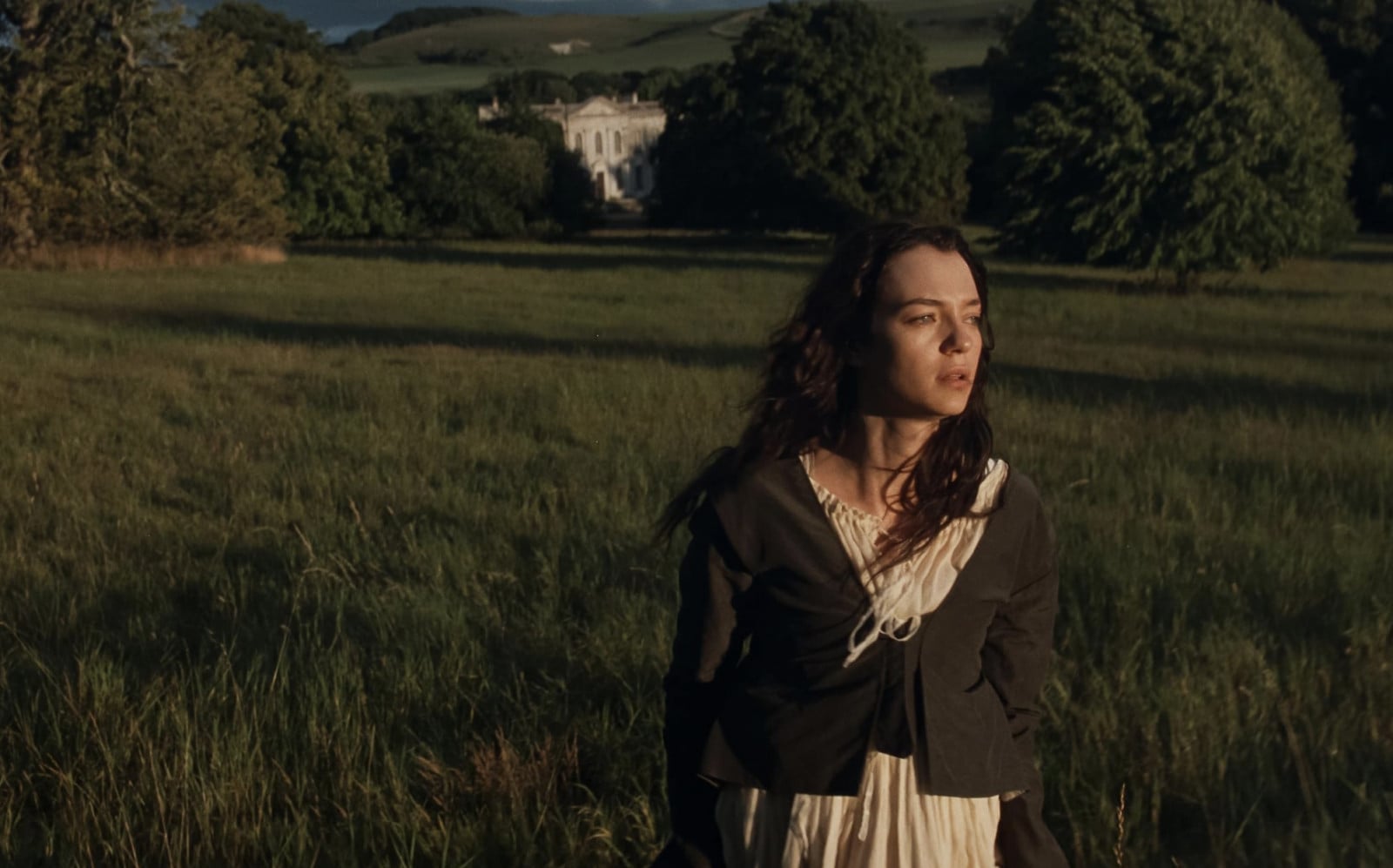 A woman in Regency-era attire stands in a sweeping meadow, a grand estate visible in the distance.