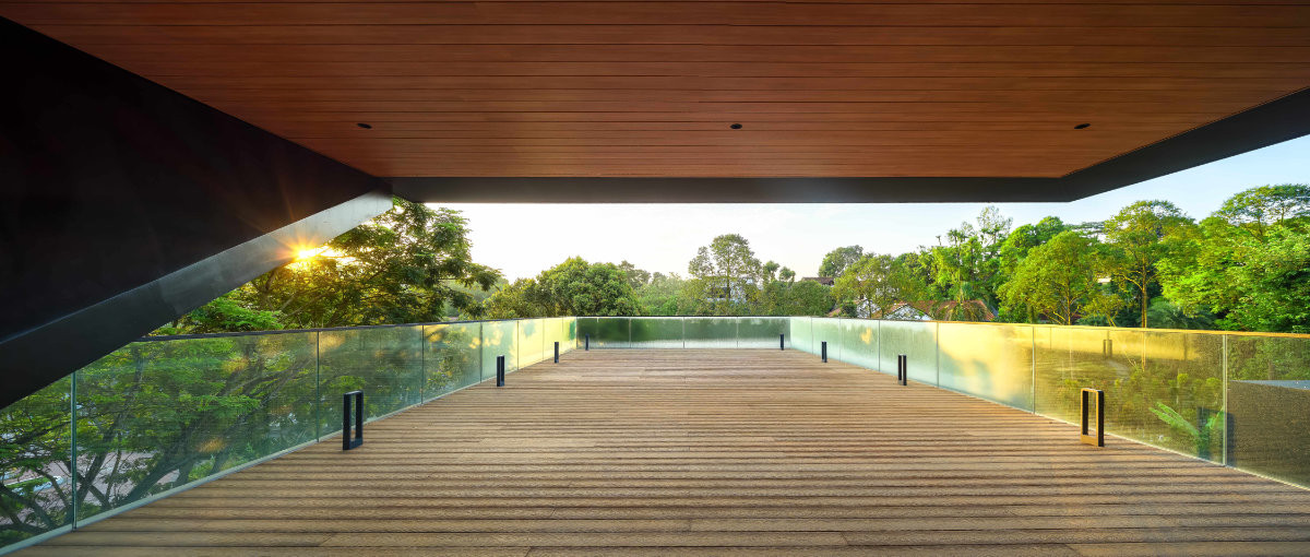 A timber-decked sky terrace sits beneath a deep cantilevered roof, its glass balustrade dissolving into the treetops at sunset.