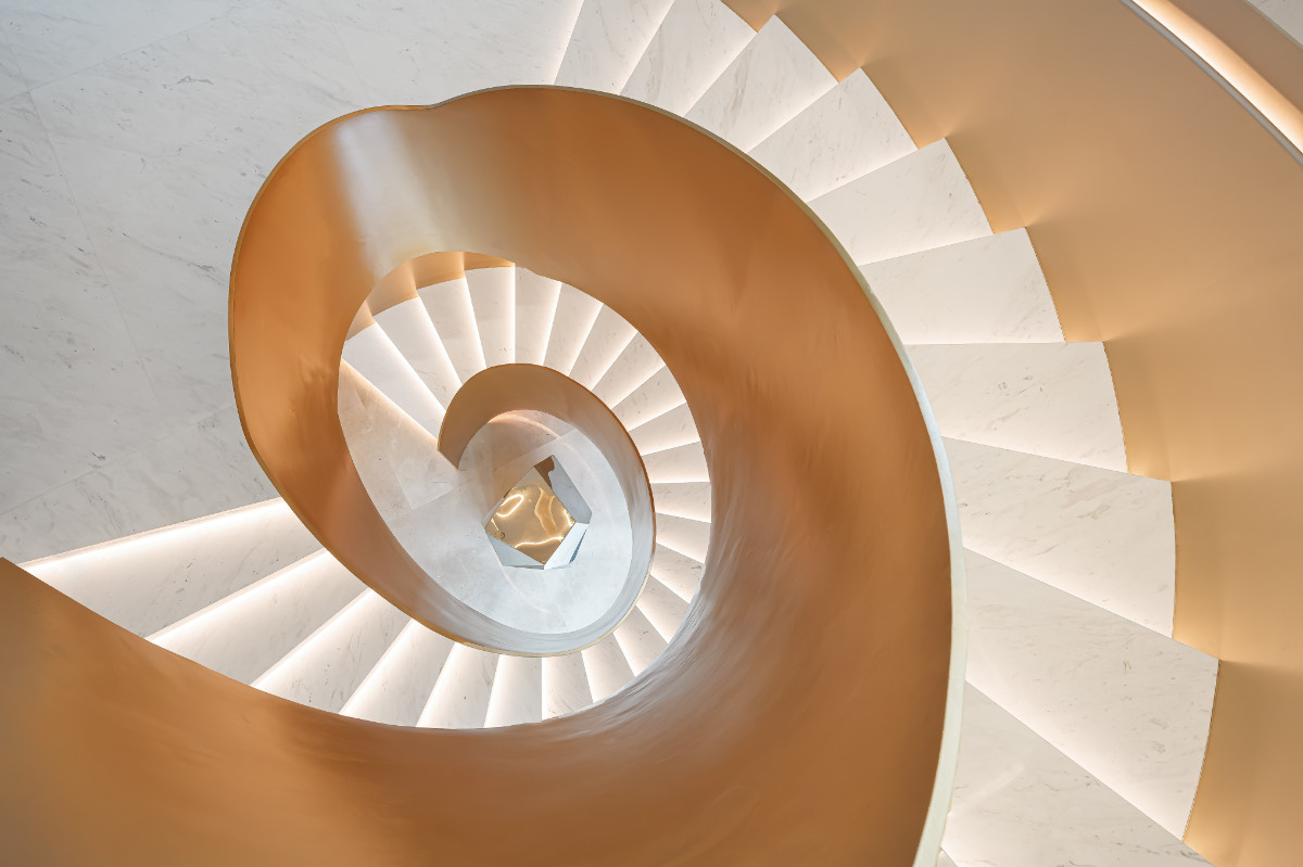 Seen from above, the spiral staircase unfurls like a ribbon, its champagne-toned casing and lit marble treads coiling down through the levels of the house