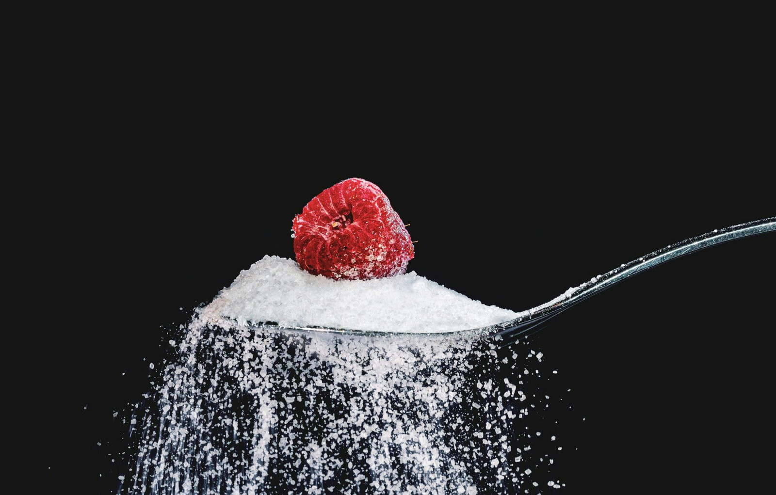 White granulated sugar, a common sweetener, spills from a spoon with a fresh raspberry.