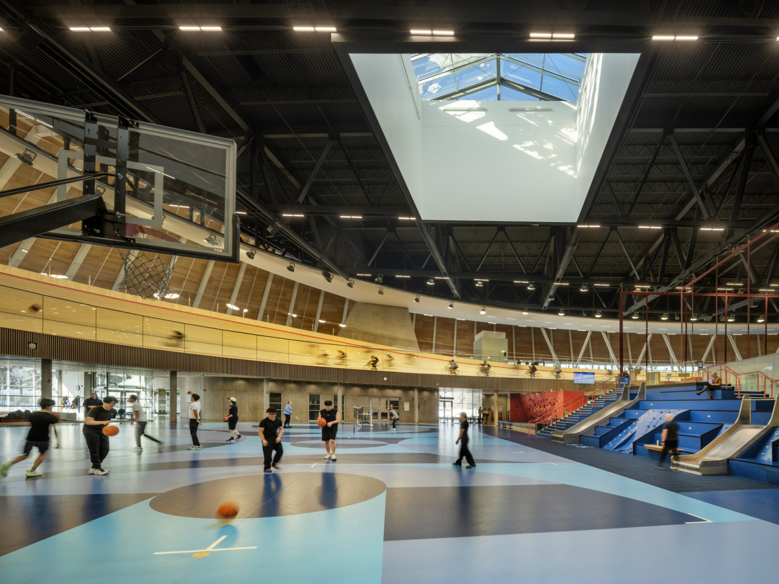 From the basketball courts on the ground floor, visitors can watch cyclists on the raised velodrome above, while a skylight brings daylight into the hall and tiered blue seating with slides lines one side. (Photo: Nic Lehoux)