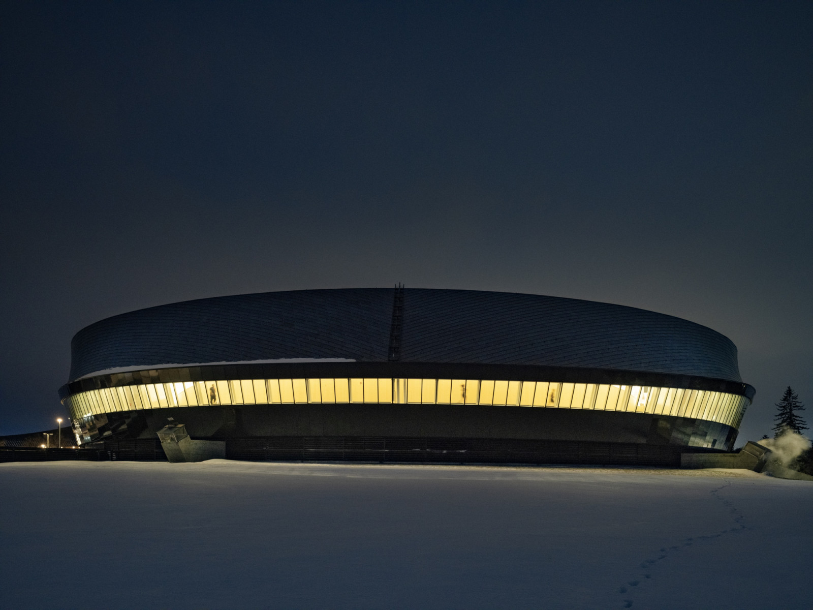On a winter night, the lit band of glazing that wraps Coronation Park Centre shows runners moving around the track inside (Photo: Nic Lehoux)
