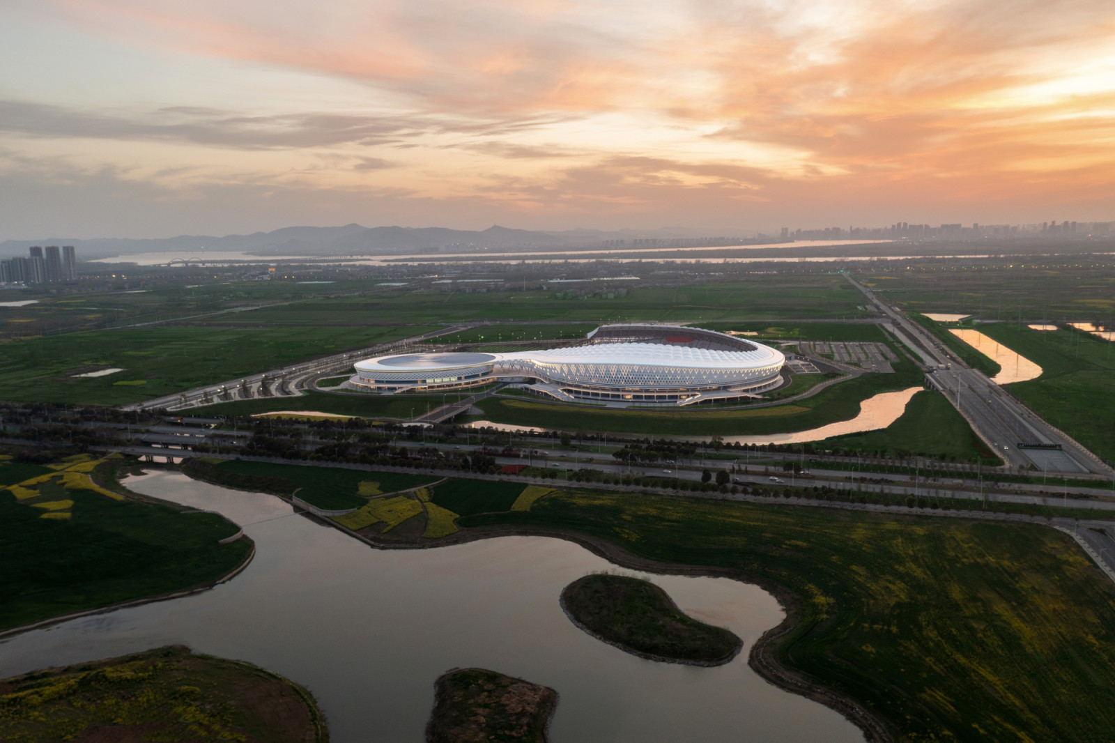 Xiangyang Olympic Center Stadium sits among green fields and waterways in Hubei Province, its white canopy linking the stadium to a circular venue beside it (Photo: Yilong Zhao)
