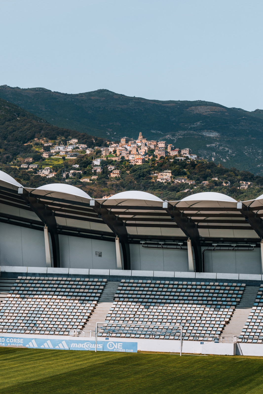 The canopy’s domed membrane bays sit above blue and white seating, framing a view of a hillside village behind the stadium. (Photo: Banzaiprod)