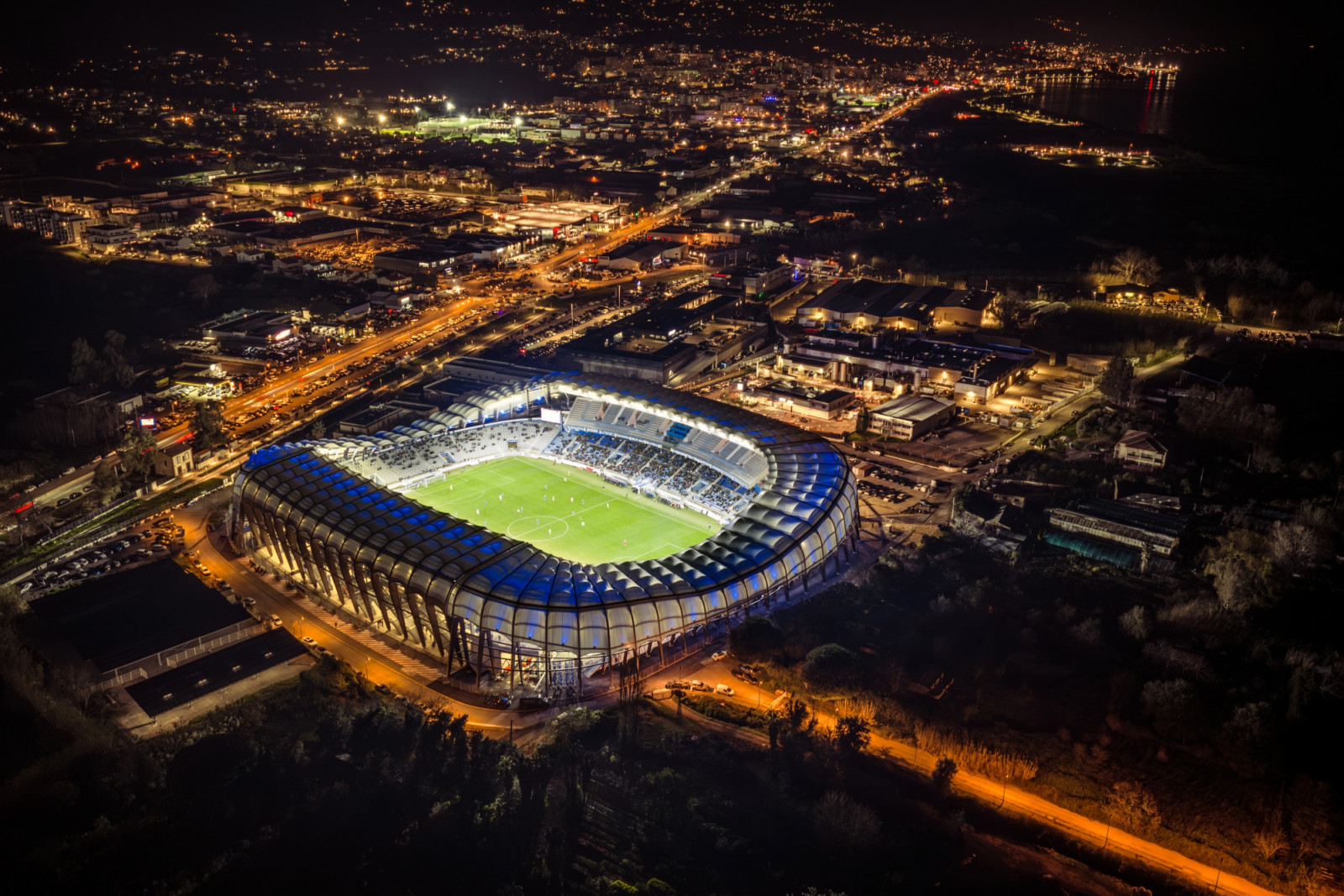 On a match night, blue lighting picks out the canopy’s rounded bays above the 16,500-seat terraces, with the lights of the surrounding town beyond. (Photo: Banzaiprod)