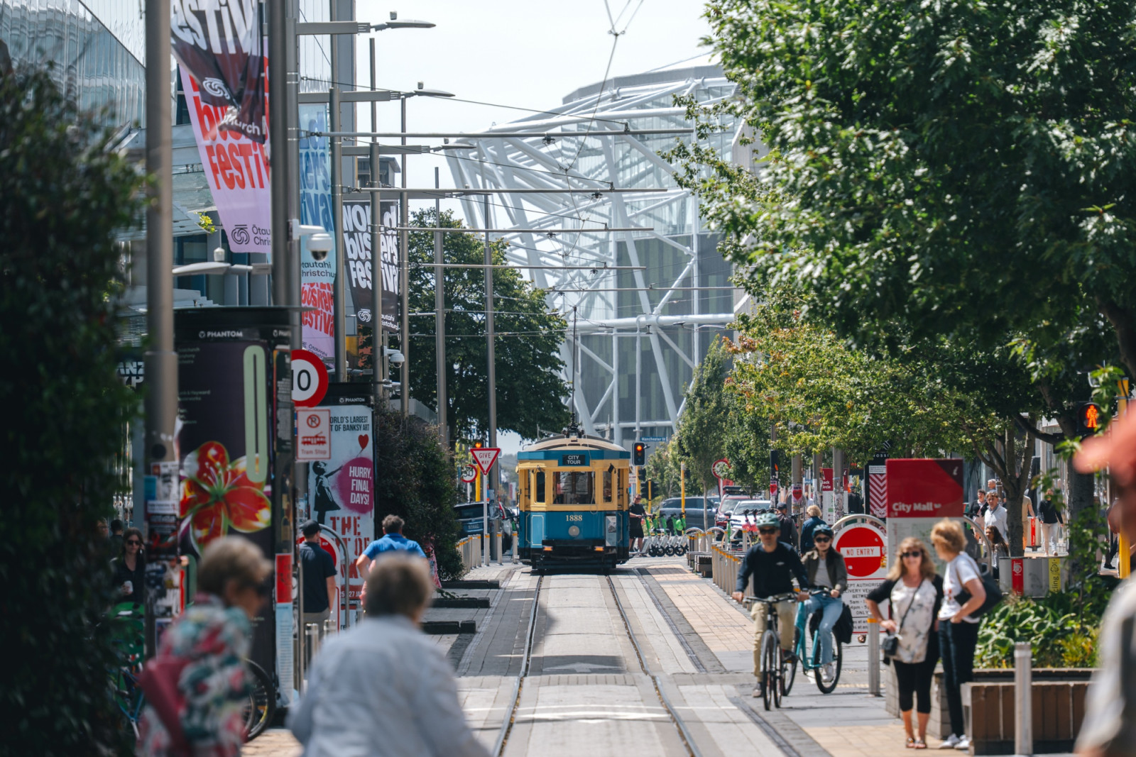 A tram travels along a city-centre street towards the stadium’s white steel framework. Te Kaha opened in April 2026 (Photo: Joel Kearns-Docker)