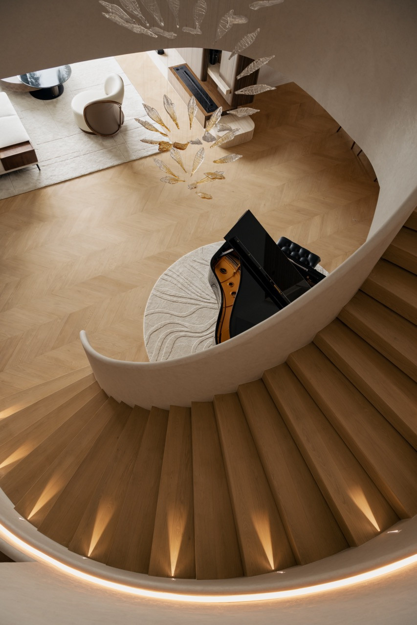 Seen from above, the staircase curves around the piano and the chevron-patterned solid wood floor, beneath a glittering chandelier