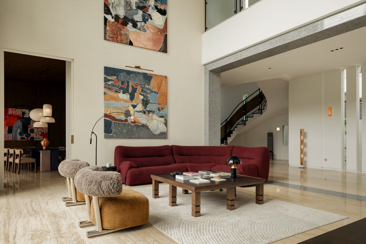 The double-height living room, where a burgundy sofa and a pair of abstract canvases sit against the marble floor, with the curved staircase visible beyond